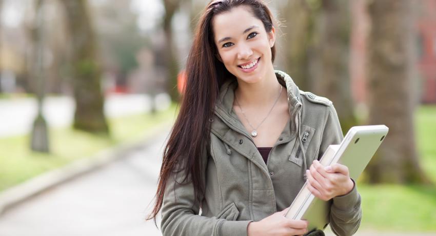 Smiling girl holding books and laptop on campus.