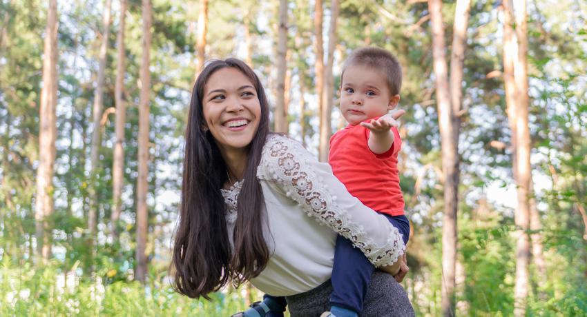 Smiling mother gives son a piggy back ride in the forest as he reaches toward the viewer. 