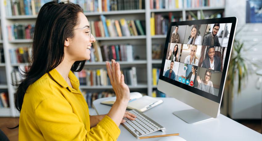 Woman with long brown hair, glasses, wearing bright yellow shirt, waving at computer screen with images of other people on screen looking back at her.